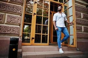 elegante uomo arabo alto modello in camicia bianca, jeans e occhiali da sole posato in una strada della città. barba attraente ragazzo arabo contro la porta d'ingresso del ristorante. foto