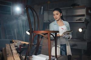 il giovane falegname asiatico di mobili di design donna utilizza un metro a nastro per misurare la sedia sul banco di lavoro nel laboratorio di falegnameria di falegnameria. foto