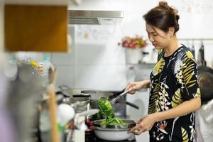 un autentico scatto reale di una donna asiatica che bolle e cucina verdure verdi per il cibo in cucina. foto