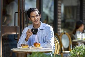 uomo asiatico sorseggiando un caffè espresso caldo mentre è seduto fuori dal bistrot del caffè in stile europeo godendosi la vita lenta con l'atmosfera mattutina nella piazza della città con pasticceria dolce foto