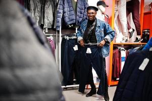 uomo afroamericano casual elegante alla giacca di jeans e berretto nero al negozio di vestiti alla ricerca di nuovi pantaloni sportivi. foto