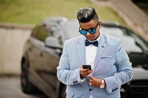 elegante uomo arabo in giacca, papillon e occhiali da sole contro un'auto suv nera. uomo d'affari ricco arabo con il telefono cellulare. foto