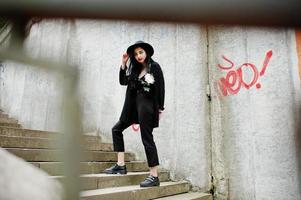 ragazza sensuale tutta in nero, labbra rosse e cappello. la donna drammatica goth tiene il fiore del crisantemo bianco contro il muro dei graffiti. foto