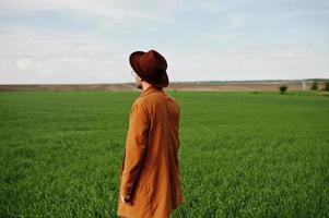 uomo alla moda con gli occhiali, giacca marrone e cappello in posa sul campo verde. foto