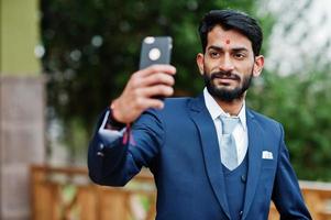 uomo d'affari indiano con barba elegante con bindi sulla fronte, indossare un abito blu posato all'aperto e fare selfie al telefono cellulare. foto