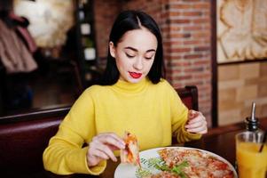 divertente ragazza bruna in maglione giallo che mangia pizza al ristorante. foto