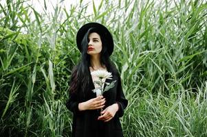 ragazza sensuale tutta in nero, labbra rosse e cappello. goth donna drammatica su canna comune tenere fiore di crisantemo bianco. foto