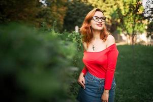 attraente donna dai capelli rossi in occhiali, indossare su camicetta rossa e gonna di jeans in posa al parco verde. foto