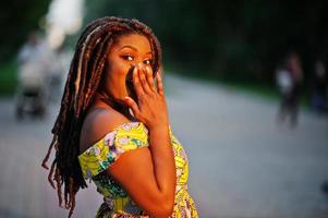 carina ragazza afroamericana di piccola altezza con i dreadlocks, indossa un vestito giallo colorato, posata al tramonto, sorpresa di emozione. foto