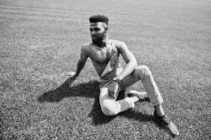 bell'uomo a torso nudo afroamericano sexy in tuta di jeans seduto all'erba verde del campo di calcio dello stadio. ritratto di uomo nero alla moda. foto