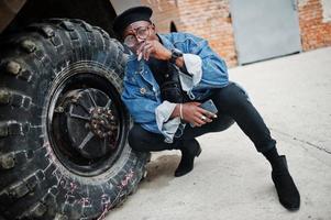 uomo afroamericano in giacca di jeans, berretto e occhiali da vista, fumando sigaro e posato contro la ruota del veicolo blindato militare btr. foto