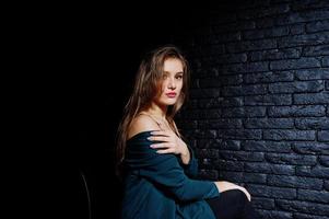 bella ragazza bruna indossa una giacca nera e verde, seduta e posando su una sedia in studio contro un muro di mattoni scuri. ritratto del modello in studio. foto