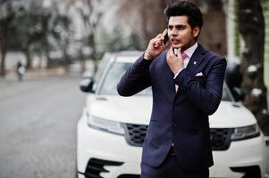 elegante modello di uomo macho indiano su abito e cravatta rosa in posa contro un'auto bianca e parlando al telefono. foto