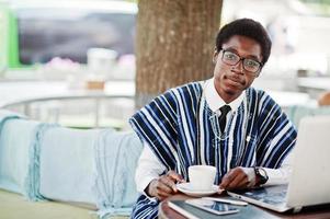 uomo africano in abiti tradizionali e occhiali seduto al caffè all'aperto, bere caffè e lavorare al computer portatile. foto