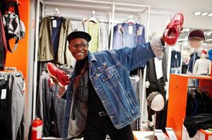 uomo afroamericano casual elegante alla giacca di jeans e berretto nero al negozio di vestiti che tiene nuove scarpe da ginnastica rosse a portata di mano. foto
