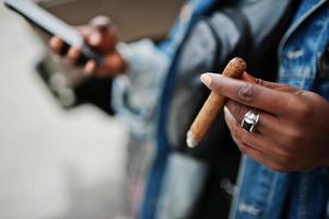 mani ravvicinate dell'uomo afroamericano in giacca di jeans, sigaro fumante con il cellulare a portata di mano. foto