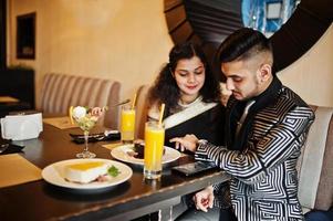 adorabile coppia indiana innamorata, indossata a saree e abito elegante, seduta al ristorante. a tavola dolci e succhi di frutta. uomo che guarda i suoi orologi. foto