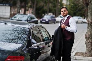 elegante uomo d'affari indiano in abbigliamento formale in piedi contro un'auto nera d'affari sulla strada della città. aprire la porta dell'auto con la chiave. foto