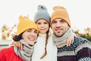 il ritratto di una bambina adorabile indossa un cappello lavorato a maglia e un maglione si trova tra i genitori, abbracciali. la bella donna indossa una sciarpa calda e un maglione che gode del tempo libero con il marito e la figlia piccola foto