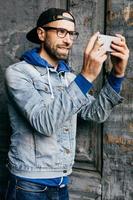 ritratto di ragazzo alla moda con la barba che indossa abiti alla moda che tiene il telefono cellulare facendo selfie essendo contento e sorridente con gioia nella fotocamera. maschio rilassato con aspetto attraente in posa a porte chiuse foto