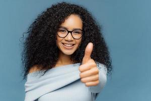 il ritratto di una donna dalla pelle scura ha il pollice in su, mostra accordo o approvazione, ha un sorriso sul viso, indossa occhiali e maglione, gesti al chiuso su sfondo blu. linguaggio del corpo e concetto ok foto