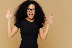 inquadratura a metà lunghezza di una donna felicissima e contenta balla e si muove positivamente, alza i palmi delle mani, esclama di felicità, indossa una maglietta nera, occhiali, isolata sul muro beige. le modelle eccitate si muovono foto