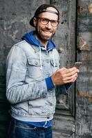 giovane uomo barbuto alla moda con berretto, camicia di jeans e occhiali che tiene lo smartphone in mano facendo foto mentre si trova vicino alla superficie della fessura. ragazzo felice hipster con gadget nelle sue mani