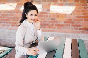 donna d'affari di successo con capelli scuri, begli occhi, pelle sana, labbra ben modellate con unghie lunghe rosse che indossano abiti formali mentre si siede al bar usando il laptop per la comunicazione online e il lavoro foto