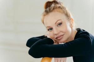 primo piano di una donna dai capelli rossi felice con il trucco, pratica il balletto in studio, si appoggia le mani sulle barre orizzontali, indossa un costume nero per la ballerina. la ballerina ha una pelle sana, si gode il riposo foto