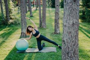 colpo all'aperto di donna attiva si sporge da parte fa esercizi di aerobica con palla fitness, vestito con top corto e leggings, posa su karemat contro alberi ed erba verde, fa sport nella natura foto