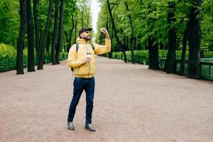 ritratto all'aperto di giovane bel turista vestito con abiti eleganti che tiene zaino e caffè da asporto in posa nella fotocamera del suo smartphone facendo selfie isolato su sfondo verde natura foto