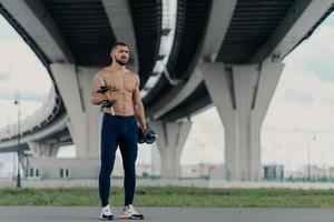 muscoloso atletico bodybuilder barbuto solleva manubri lavora sui bicipiti ha un duro allenamento vicino al ponte vestito con pantaloni sportivi e scarpe da ginnastica, essendo motivato, ama lo sport. concetto di stile di vita sano foto