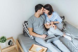 adorabili coppie romantiche che si baciano, si guardano mentre si siedono sul letto, usano il computer portatile per guardare film, bevono caffè, si divertono insieme e si rilassano in camera da letto. concetto di relazione foto