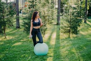 la foto all'aperto di una giovane donna sportiva tiene una bottiglia d'acqua fresca, ascolta la musica negli auricolari, tiene uno smartphone moderno, si appoggia al ginocchio sulla palla fitness, si gode la playlist preferita, posa nella foresta verde