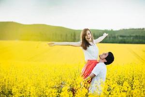 mi sento libera gioiosa giovane donna alza le mani essendo sulle mani dell'uomo, posa insieme sul campo di fiori gialli durante il soleggiato clima estivo. le coppie romantiche si divertono all'aperto. concetto di relazioni. foto