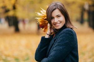 bella donna europea dai capelli scuri con espressione soddisfatta, vestita con un cappotto caldo, tiene fogliame, posa su sfondo giallo sfocato posa nel parco, posa alla telecamera, esprime felicità foto