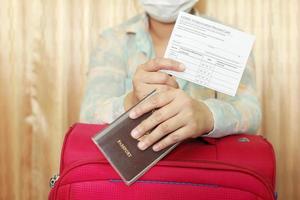 donna in abiti da viaggio che indossa una maschera medica che mostra la tessera di vaccinazione covid-19 e il passaporto e la valigia rosa. concetto di viaggio con vaccinazione. foto
