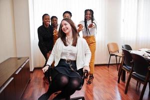 cinque donne d'affari multirazziali in piedi in ufficio e rotolare donna sulla sedia. gruppo eterogeneo di dipendenti in abiti formali che si divertono. foto