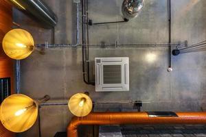 alzando lo sguardo sul soffitto in cemento grigio con faretti alogeni e lampade edison in un ufficio loft con aria condizionata e tubo di ventilazione arancione foto