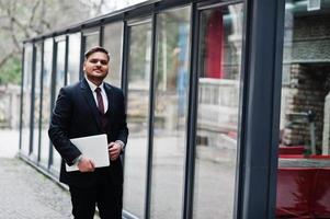 elegante uomo d'affari indiano in abbigliamento formale con laptop sulle mani in piedi contro le finestre nel business center. foto