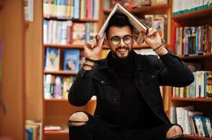 studente arabo alto e intelligente, indossa una giacca di jeans nera e occhiali da vista, seduto in biblioteca e coprendo la testa con un libro. foto