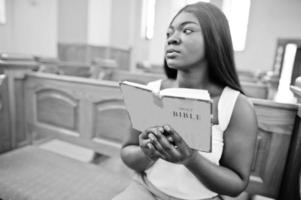 donna afroamericana che prega nella chiesa. i credenti meditano nella cattedrale e nel tempo spirituale della preghiera. ragazza afro tiene la Sacra Bibbia mentre è seduto su una panchina. foto