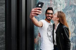 bella coppia multirazziale in posa contro il muro e facendo selfie insieme. ragazza europea bacia l'uomo arabo sulla guancia. foto