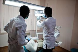 paziente uomo afroamericano in poltrona odontoiatrica. studio dentistico e concetto di pratica medica. dentista professionista che aiuta il suo paziente in odontoiatria medica. indicando la radiografia dei denti. foto