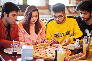 gruppo di amici asiatici che mangiano pizza durante la festa in pizzeria. felici indiani che si divertono insieme, mangiano cibo italiano e si siedono sul divano. foto