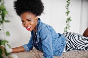 attraente donna afroamericana con capelli afro su gonna e giacca di jeans, posata in una stanza bianca sull'altalena. modello nero alla moda. foto