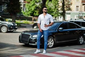 elegante uomo arabo alto modello in camicia bianca, jeans e occhiali da sole posato in una strada della città. barba attraente ragazzo arabo con una tazza di caffè contro l'auto nera di affari. foto