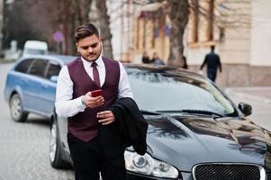elegante uomo d'affari indiano in abbigliamento formale con il telefono cellulare in piedi contro un'auto nera d'affari sulla strada della città. foto