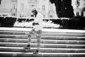 elegante uomo afroamericano in camicia bianca e pantaloni colorati con cappello e occhiali in posa all'aperto. ragazzo nero modello alla moda. foto