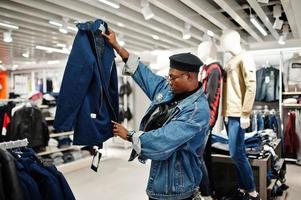 uomo afroamericano casual elegante alla giacca di jeans e berretto nero al negozio di vestiti che guarda la nuova giacca. foto